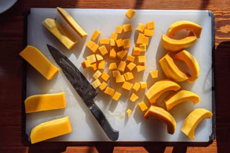 A knife sits on a white plastic cutting board with pieces of a butternut squash cut up around it.